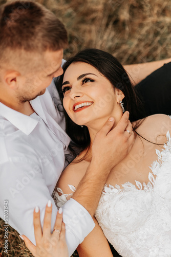 Groom and smiling bride lie on dry grass and hug, woman in white wedding dress. Beautiful autumn wedding photo. The groom aims at her forehead