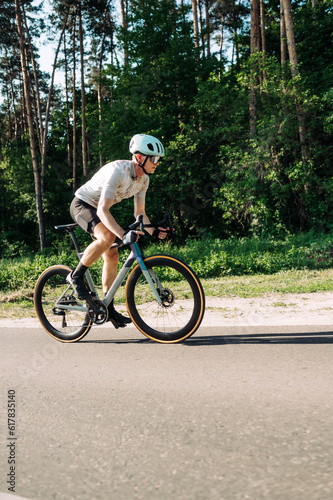 Fototapeta Naklejka Na Ścianę i Meble -  Professional cyclist in sports equipment trains on a road bike outside the city, rides on the road against the background of the forest.