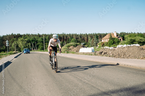 A male cyclist rides a road bike on an asphalt road outside the city on a sunny day, wearing sports equipment.