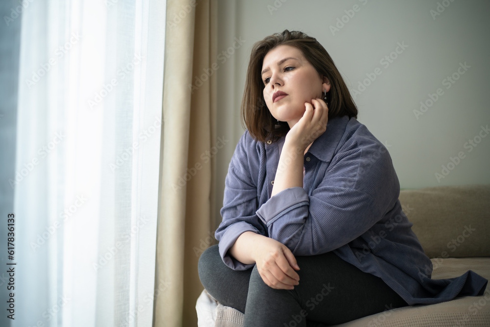 Sad plump or overweight young girl looks out window portrait. Problems of loneliness and mental health plus size people in society. Sad plump or overweight young girl looks out window portrait. Problems of loneliness and mental health plus size people in society.