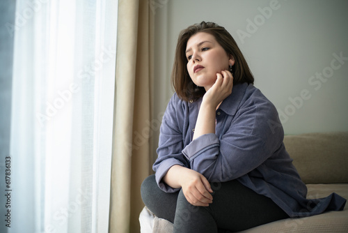 Sad plump or overweight young girl looks out window portrait. Problems of loneliness and mental health plus size people in society. photo