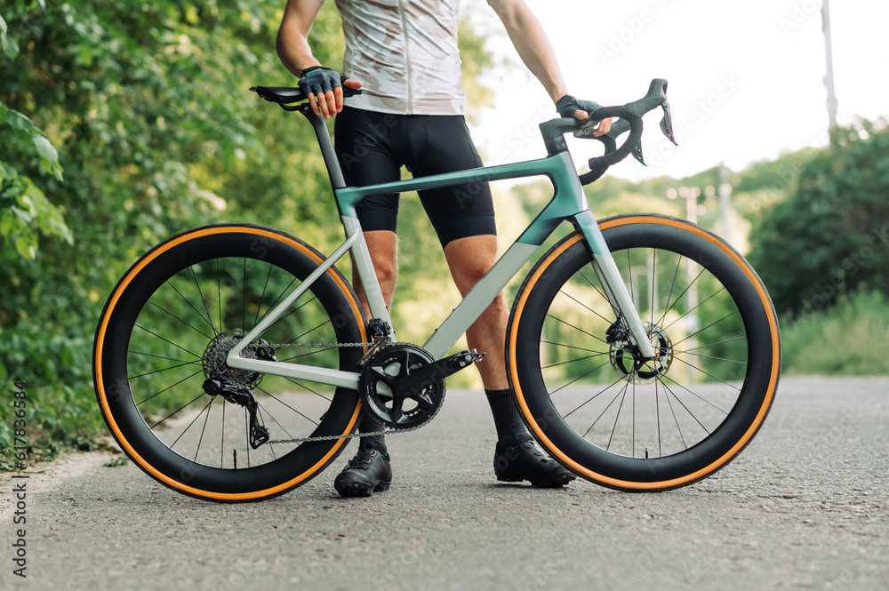 A male cyclist in gear stands with a professional road bike on an asphalt road outside the city. Close up photo. Sport concept A male cyclist in gear stands with a professional road bike on an asphalt road outside the city. Close up photo. Sport concept