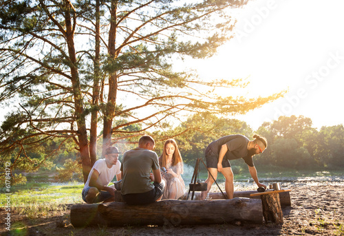 Smiling guys and girls are resting in the summer on the banks of the river with a tent. Cooking food on a fire. Hiking. Beautiful summer nature  landscape. Copy space for text