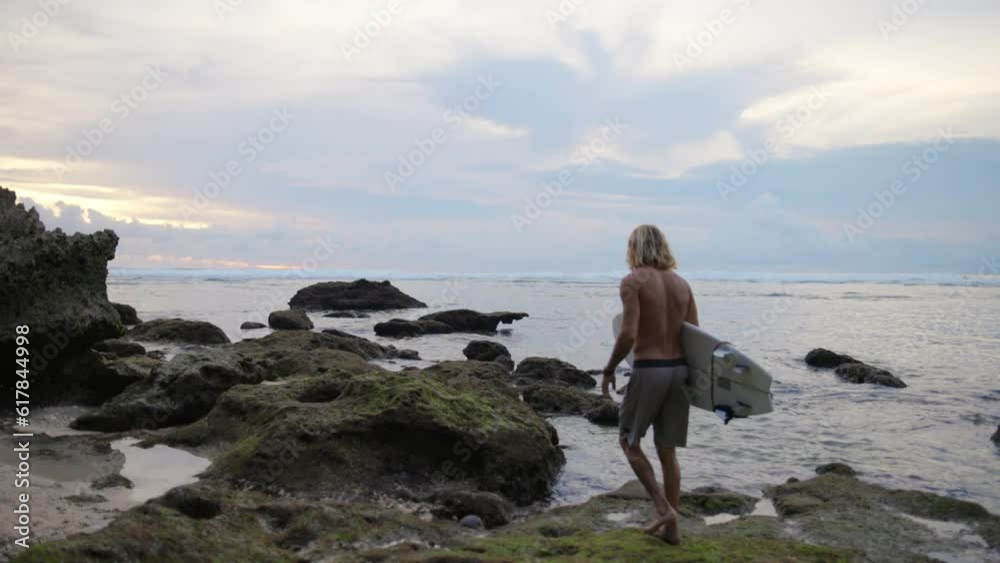 Rear view of sporty male surfer walking along rocky seashore in early morning looking for place to surf, point to surf. Surfing in early morning at sea. Man with surfboard walks along rocky shore. Rear view of sporty male surfer walking along rocky seashore in early morning looking for place to surf, point to surf. Surfing in early morning at sea. Man with surfboard walks along rocky shore.