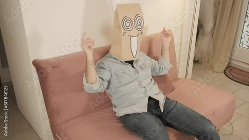A man wearing a breadbag face, sitting on the sofa at home, looking stoned (being drunk or under the influence of a drug).
