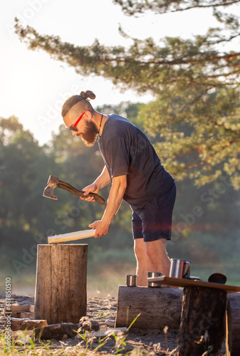 A stylish brutal man with a beard and black glasses is chopping wood on a campsite to kindle a fire. Outdoor recreation in summer. Survival, outdoor