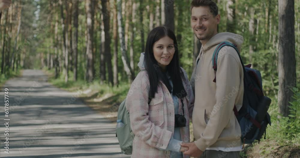Joyful young people couple with backpacks standing in forest holding hands smiling looking at camera. Summertime and vacation concept. Joyful young people couple with backpacks standing in forest holding hands smiling looking at camera. Summertime and vacation concept.