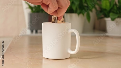 A man mixes sugar with a teaspoon in a white teacup with a tea bag and hot water to make himself tea in the kitchen. Break.
