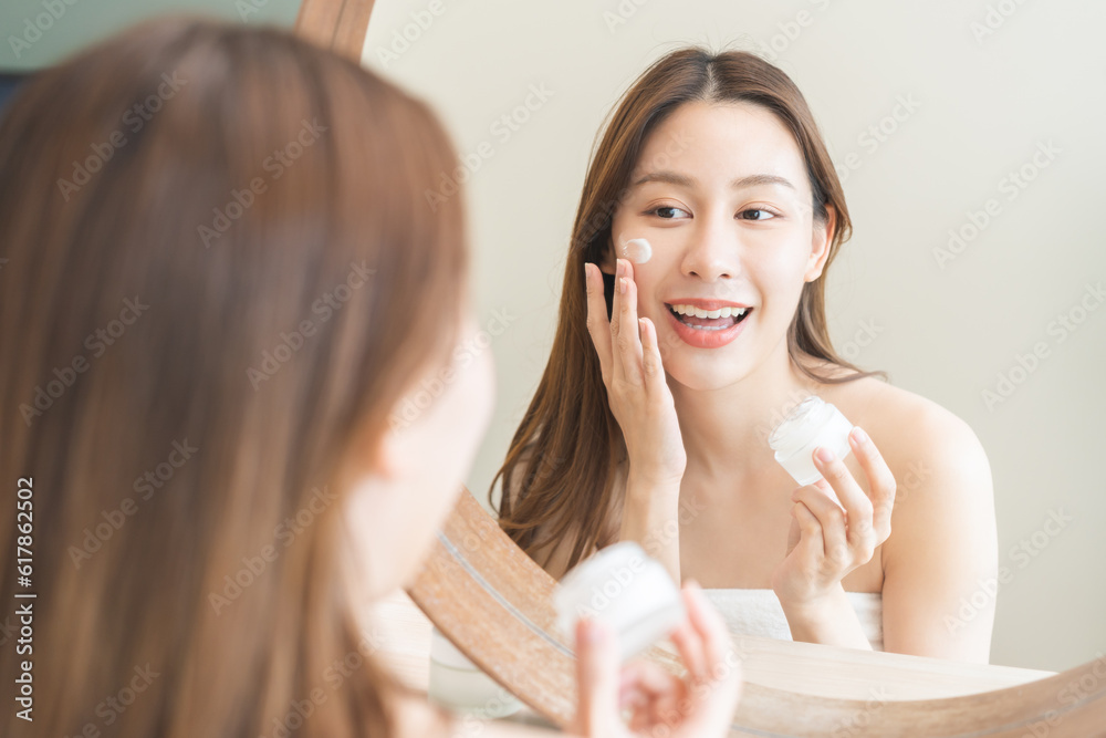 Facial beauty skin care, smile of pretty asian young woman in bathrobe looking at mirror, hand applying moisturizer lotion on her face, holding jar of skin cream before makeup cosmetic routine at home Facial beauty skin care, smile of pretty asian young woman in bathrobe looking at mirror, hand applying moisturizer lotion on her face, holding jar of skin cream before makeup cosmetic routine at home