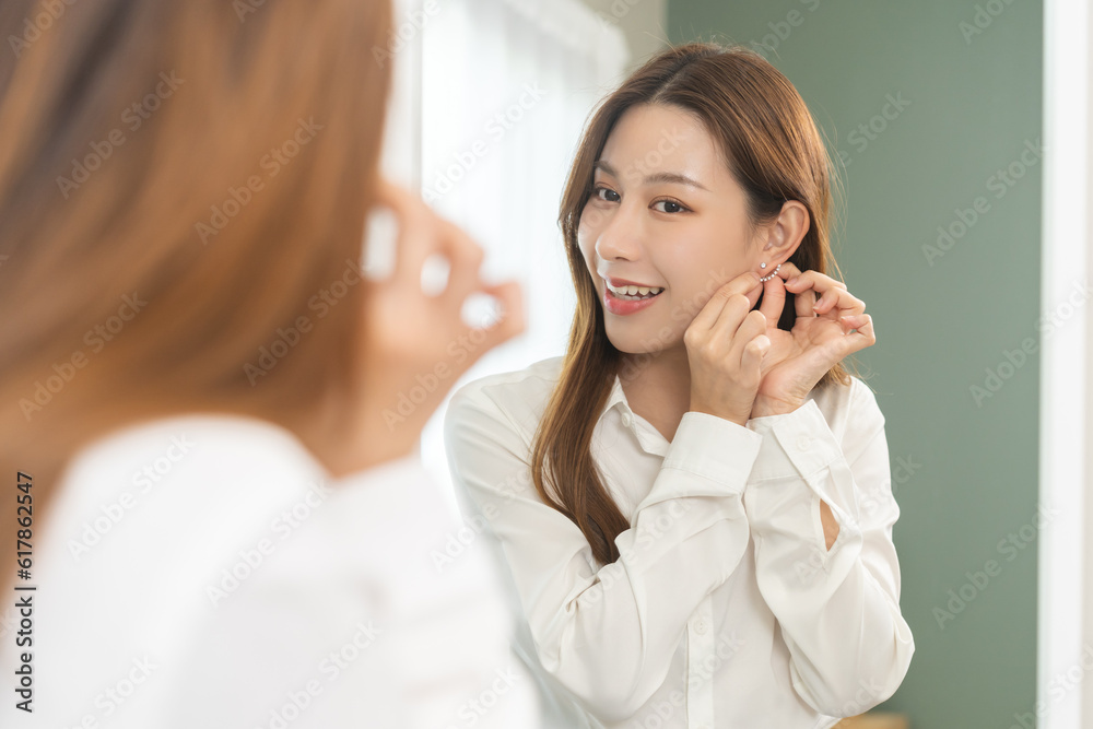 Get dress, pretty asian young woman, businesswoman standing putting earring or jewelry, wearing white shirt formal getting dressed getting ready before go to work looking reflection the mirror at home Get dress, pretty asian young woman, businesswoman standing putting earring or jewelry, wearing white shirt formal getting dressed getting ready before go to work looking reflection the mirror at home