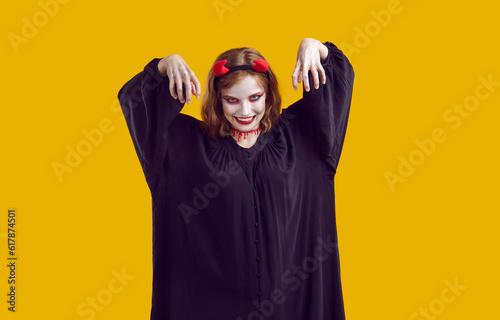 Stale corpse rises from the grave to participate in Halloween party. Woman in black robe, makeup, devil horns, bloody trail on her neck scares audience with her hands up. Isolated on yellow. © Studio Romantic