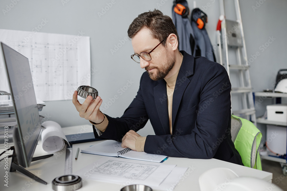 Portrait of bearded mechanical engineer holding machine part at workplace while reviewing blueprints Portrait of bearded mechanical engineer holding machine part at workplace while reviewing blueprints