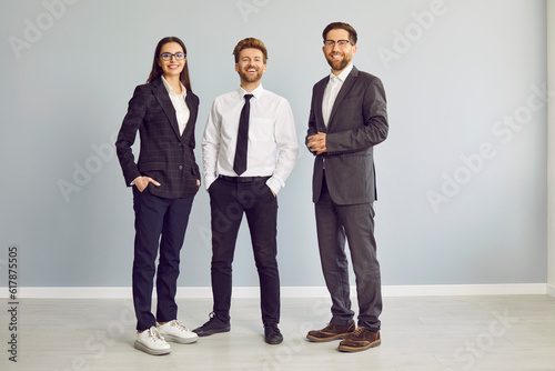 Full length portrait of a group of three young cheerful business people in suits standing confidently, looking at the camera and smiling isolated on a studio grey background. Team work concept. photo