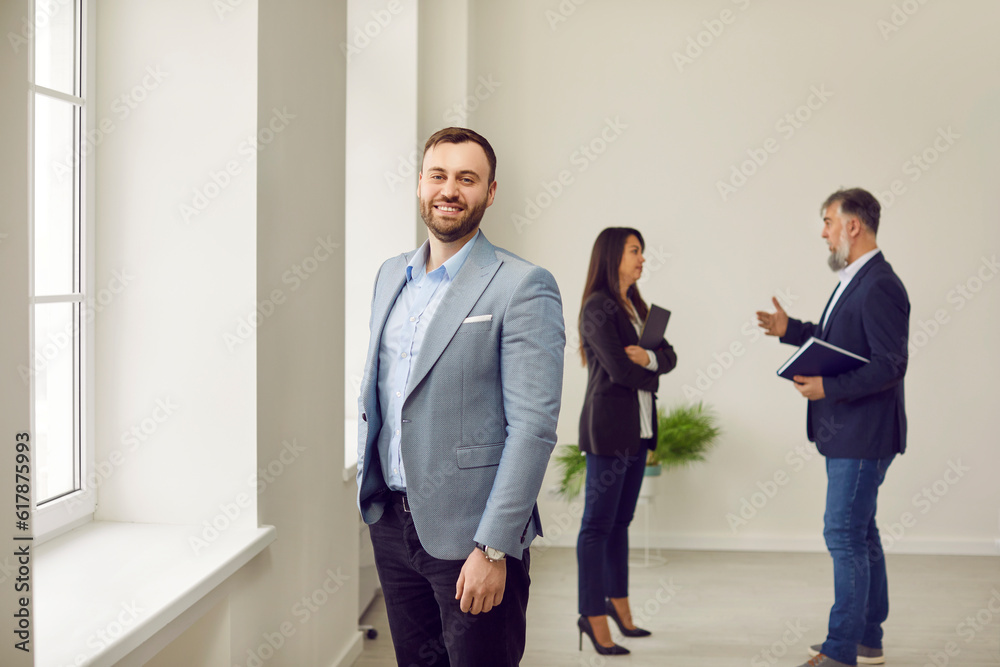 Portrait of happy smiling man looking at camera with realtor showing and discussing new apartment with his wife on background. Successful family ready to become homeowners. Real estate market concept Portrait of happy smiling man looking at camera with realtor showing and discussing new apartment with his wife on background. Successful family ready to become homeowners. Real estate market concept