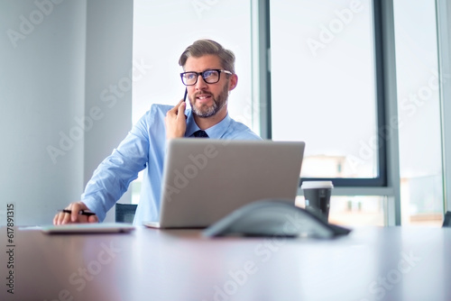 Mid aged professional male sitting in a modern office and having a business call