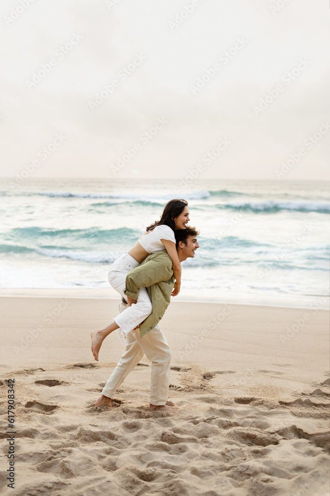 Full length shot of happy young man giving piggyback ride to his girlfriend on beach, enjoying date on coastline Full length shot of happy young man giving piggyback ride to his girlfriend on beach, enjoying date on coastline