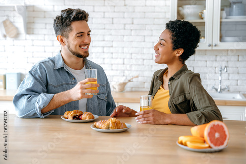 Happy couple in love, an african american woman and a caucasian man, sit at home in the kitchen, eat croissants for breakfast and drinks juice, enjoy a joint morning together, smile