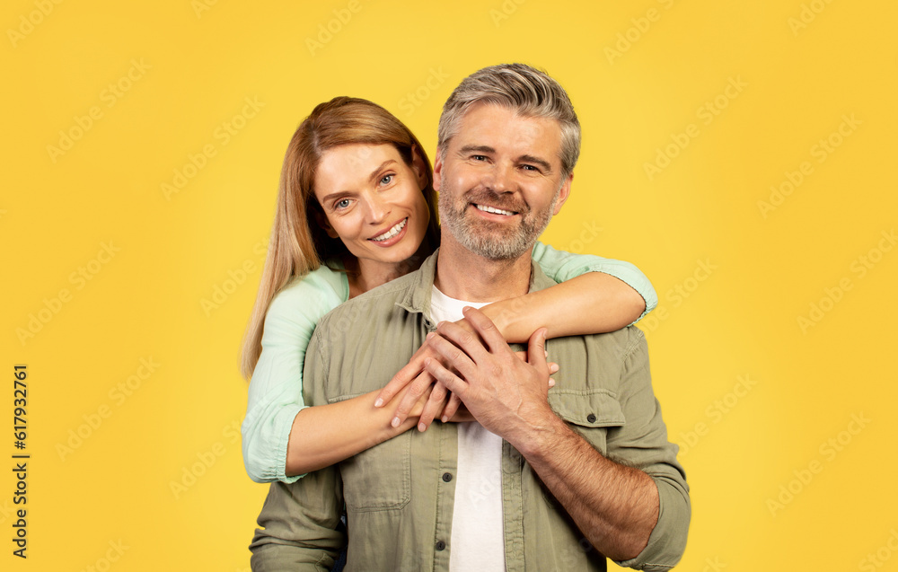 Fototapeta premium Portrait of happy middle aged woman hugging her husband from behind, posing on yellow background and smiling at camera