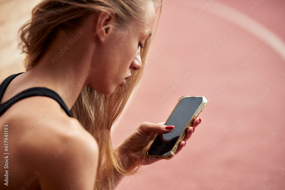 Young athlete girl is having break after a workout in the stadium. A slender attractive blonde in a black tracksuit sitting in a relaxed pose with a smartphone. Sports and recreation. Top view. Young athlete girl is having break after a workout in the stadium. A slender attractive blonde in a black tracksuit sitting in a relaxed pose with a smartphone. Sports and recreation. Top view.