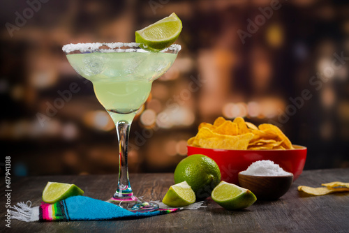 Margarita cocktail with sliced and whole limes, nachos and salt on wooden bar table. Mexican party background