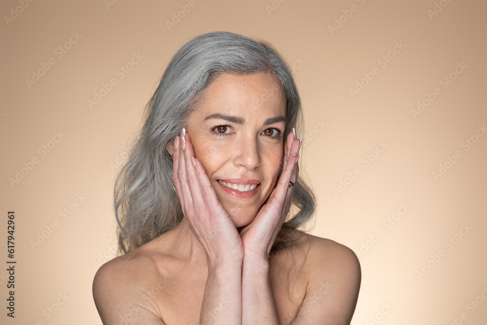 Portrait of sensual senior grey-haired woman touching her face and smiling at camera, posing over beige background