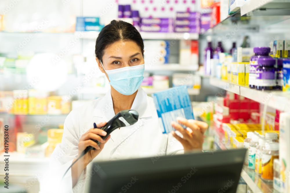 Portrait of female pharmacist in medical mask working at the cash register in pharmacy - scans barcode on the medicine package