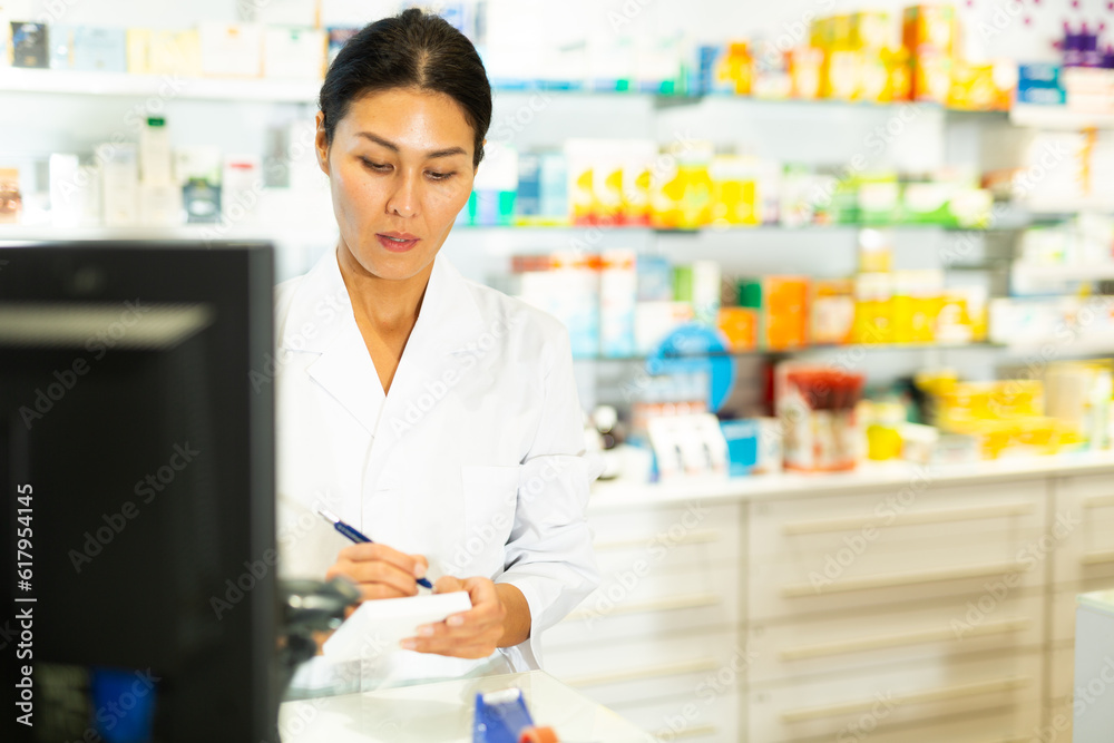Asian woman doctor standing at counter in drugstore and writing recipe.