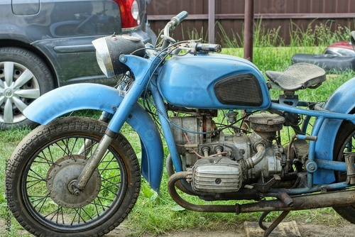 one old rusty iron classical heavy big comfortable blue dirty made in the ussr retro motorcycle stands on green grass near the car on the street during the day