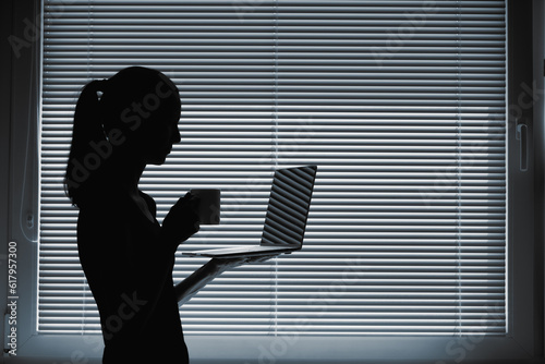 Female silhouette at window drinking coffee and looking at computer 