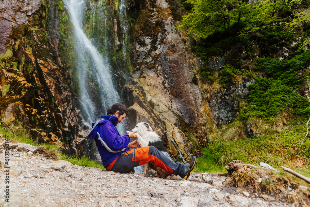 white man hiker playing with his dog in a break during a mountain path near a waterfall. travel with pet sport and adventure