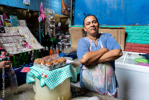 Adult Latina woman, soda vendor, arms folded watching camera in her stretch of the market in Nicaragua
