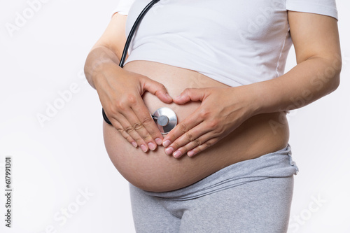 Close-up hands of a pregnant woman, expectant gravid mother forming a heart shape on her belly, listening to the heartbeat of her baby in womb with medical stethoscope, isolated on white background