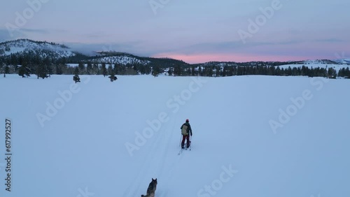 Man cross country skiing up mountain slope of Mammoth Mountain in California on snowy winter evening with German Shepherd dog running alongside