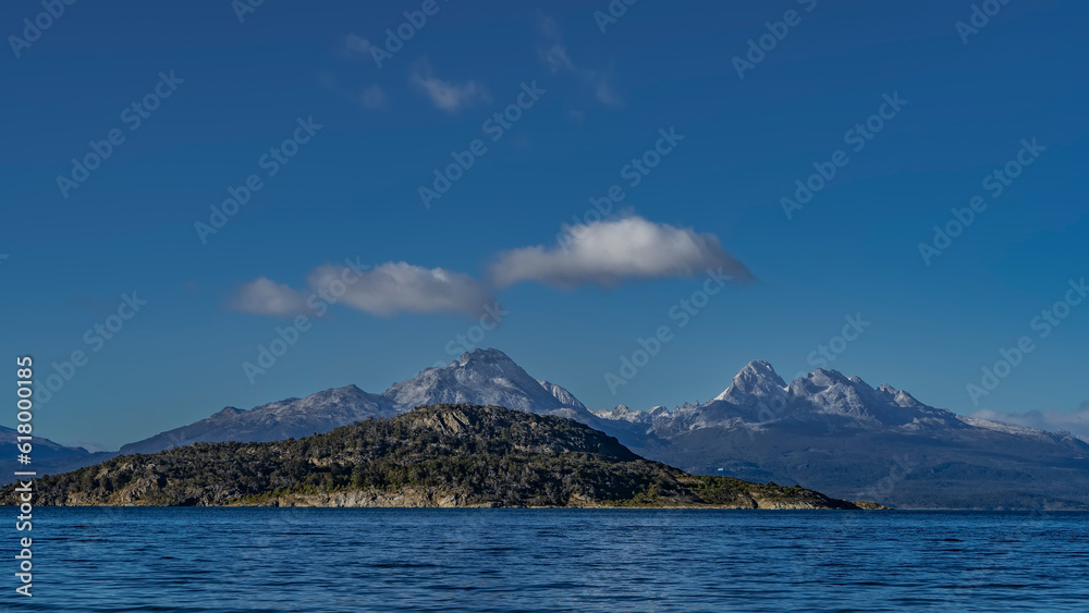 Naklejka premium A picturesque mountain range of the Andes against a background of blue sky and clouds. In the foreground is the blue water of Ensenada Bay. Argentina. Tierra del Fuego National Park. Ushuaia.