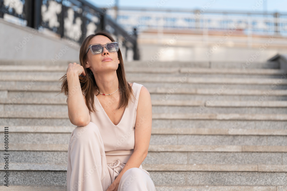 Fototapeta premium Summer urban portrait of a young woman who sits on the steps in the street.