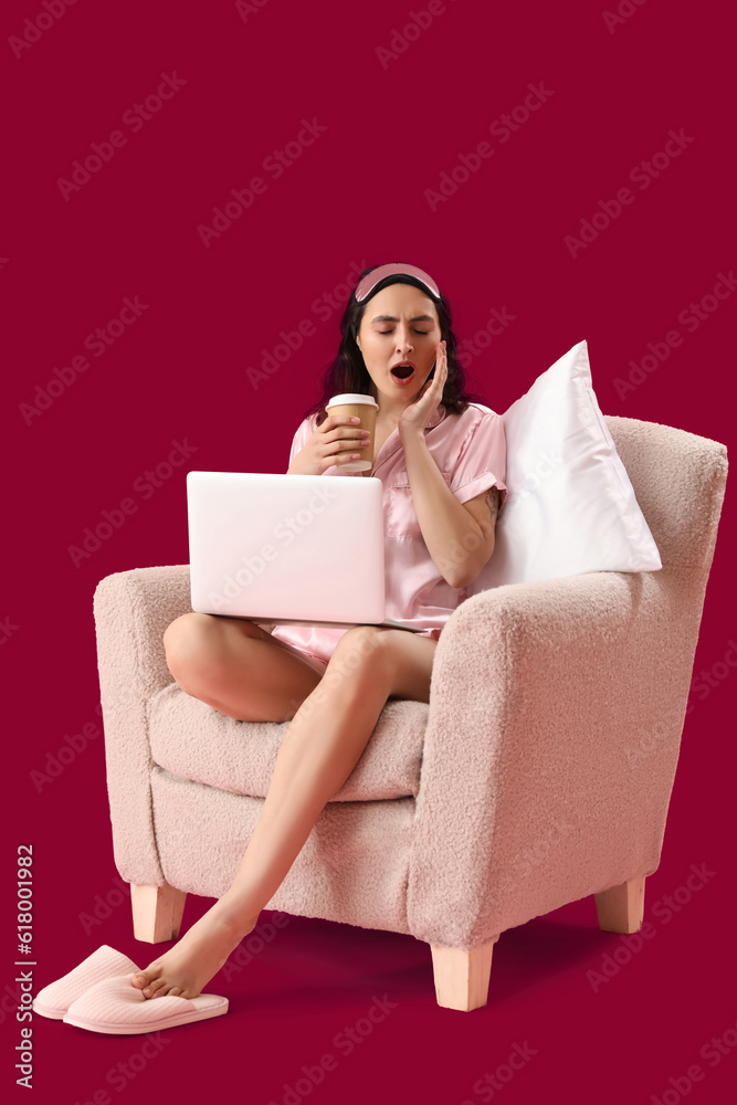 Sleepy young woman with cup of coffee using laptop in armchair on pink background Sleepy young woman with cup of coffee using laptop in armchair on pink background