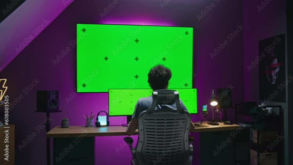 Young editor, 3D designer works at home office, edits film, develops video game, creates design for website. Teenager sits in front of multi-monitor computer and big digital screen with chromakey.