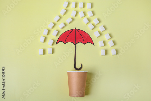 A paper cup is covered with an umbrella from sugar rain on a yellow background. The concept of reducing consumption or giving up sugar. Tea or coffee is healthy without added sugar. Flat lay