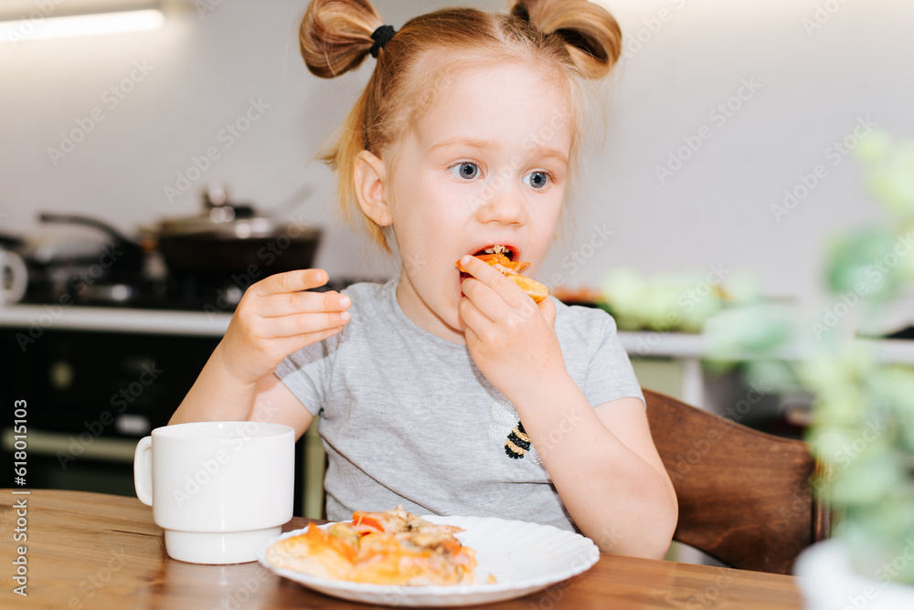 Funny little caucasian girl eating fast food while sitting at the table in the kitchen, indoors Funny little caucasian girl eating fast food while sitting at the table in the kitchen, indoors