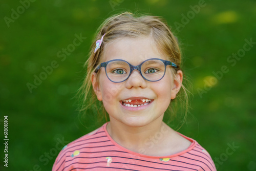 Portrait of a cute preschool girl with eye glasses and big teeth gap outdoors in park. Happy funny child on sunny summer day. Kid loosing milk tooth