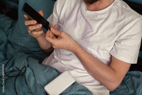 Serious Caucasian young man connect his mobile phone to a power bank while lying on his bed in the morning.