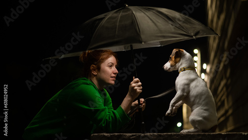 Red-haired woman and dog jack russell terrier under an umbrella in the dark. 