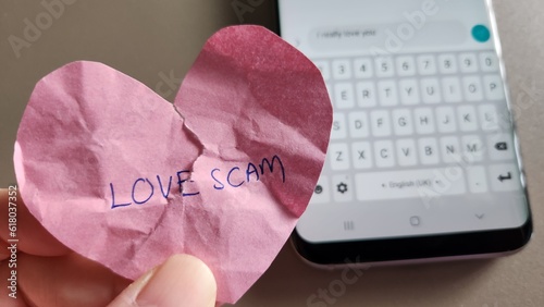 Love scam concept. A broken heart victim holding a crumpled heart with the words Love Scam written. Off focus background with a mobile phone message to the scammer  photo