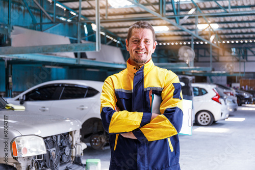 Young male mechanic in blue-yellow uniform Arms crossed and smiling at the camera with a garage in the background of cars waiting for repairs.