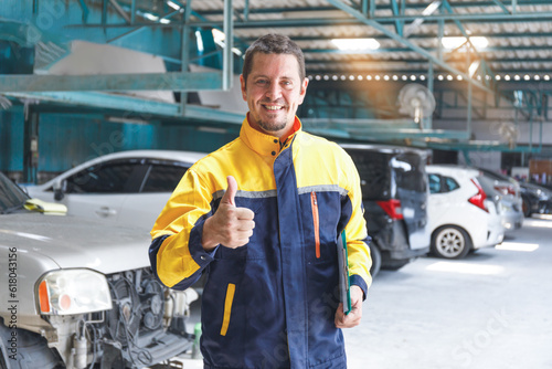 A young mechanic in a blue-yellow uniform. The mechanic stands with a thumbs up with a garage in the background of cars.