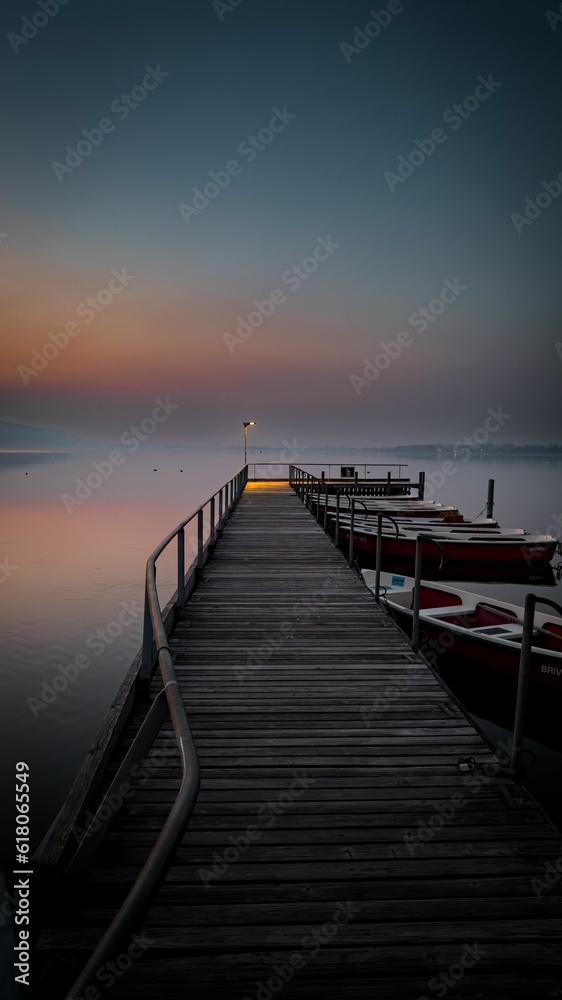 Wooden dock jutting out into a tranquil lake, with a variety of boats moored at its edges at sunset