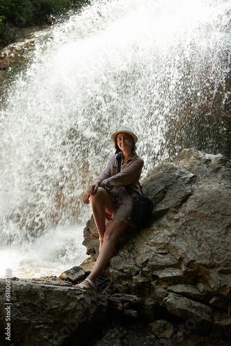 A woman sits on a rock and looks at a waterfall in the forest  a woman in a hat  a vacation in nature