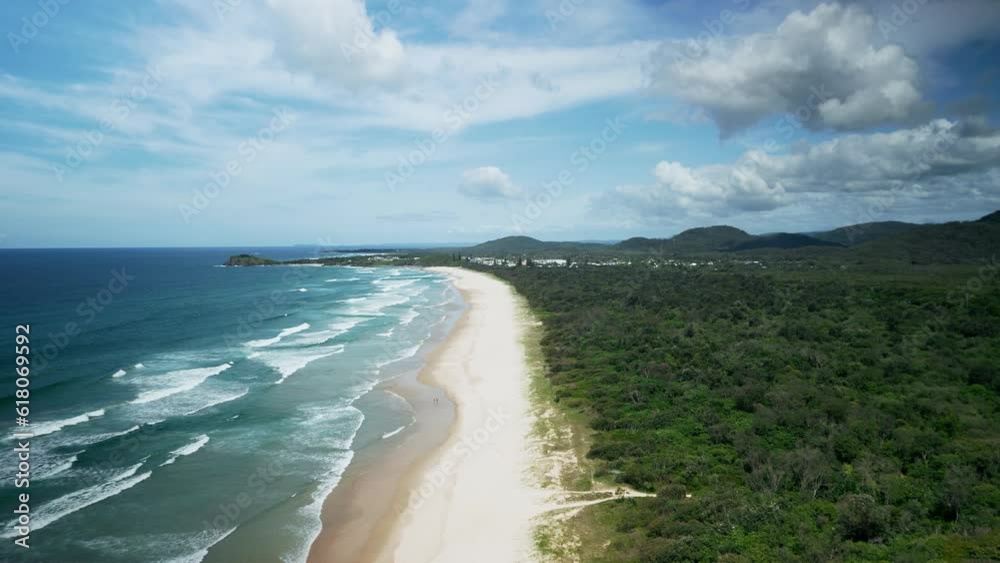 Aerial of the foamy waves of the ocean alongside the sandy shore and the green forest in Australia Aerial of the foamy waves of the ocean alongside the sandy shore and the green forest in Australia