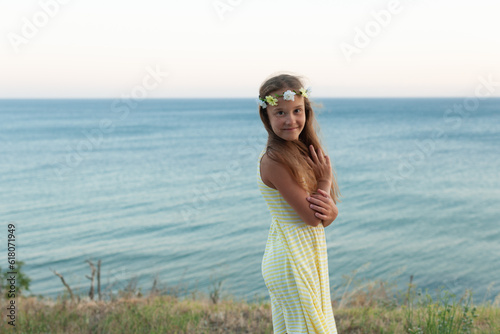 a cute caucasian girl on the beach by the sea. wearing a yellow dress with a wreath on her head. looks ahead