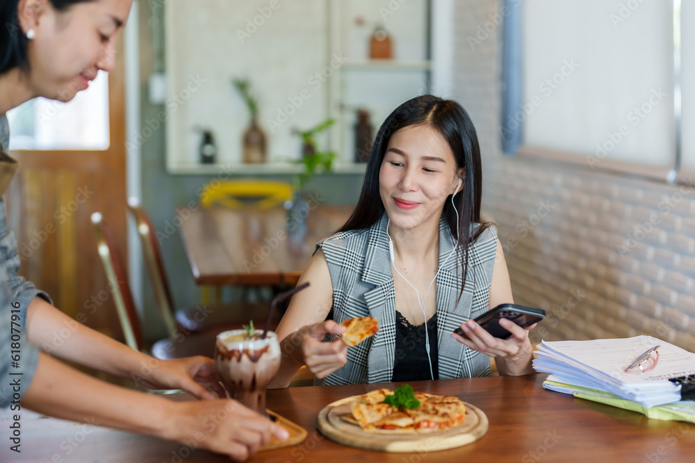 Asian woman sitting on with colleague, consult about work that was sitting in bakery cafe, plug headphones in ears sit listen music relieve stress, waitress carries ordered food places it on table.